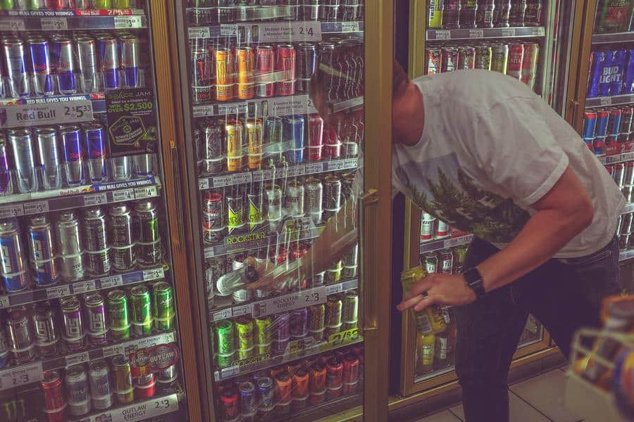 Guy in white tee picking a can of energy drink from a fridge cooler. 
