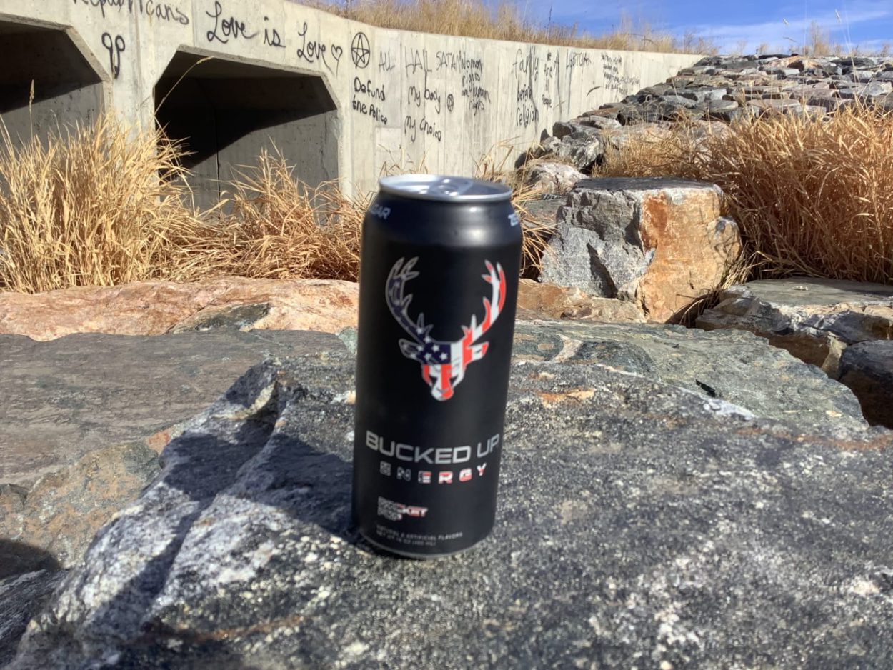 A can of Bucked Up placed on a rock with a birdge behind it
