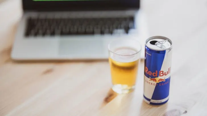 A can of Red Bull set beside a glass filled with Red Bull and a laptop