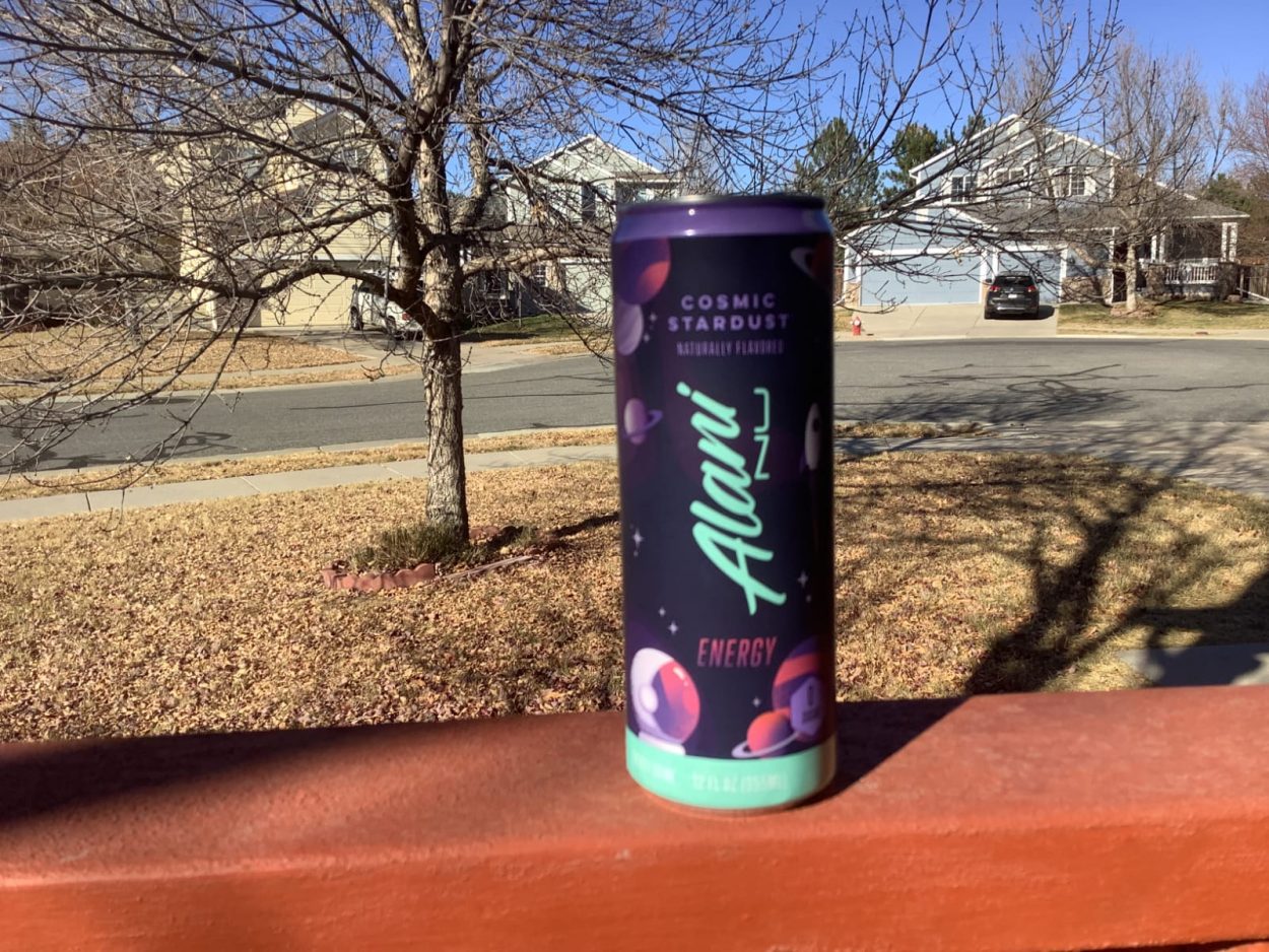 A can of Alani Nu with a tree and house with a car behind it