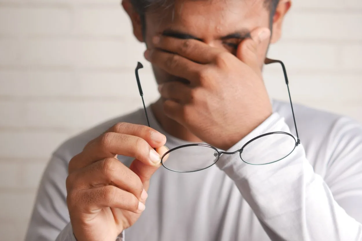 A man in white t shirt rubbing his eyes while holding his glasses